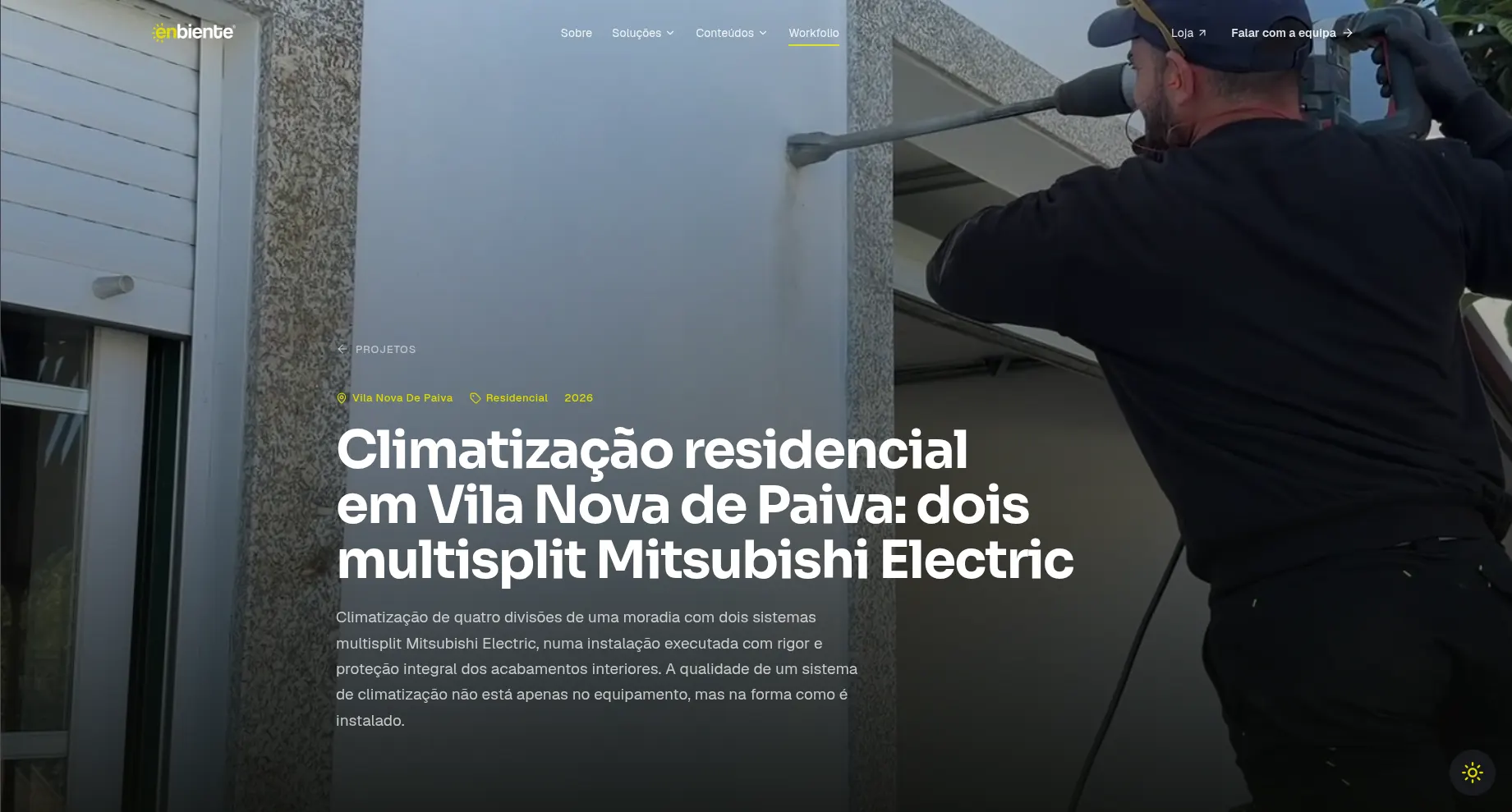 Uma página de projeto no workfolio da Enbiente, com fotografia de fundo a toda a largura de um técnico a furar uma parede exterior. Por cima, a etiqueta de local «Vila Nova De Paiva», a categoria «Residencial», o ano 2026, o título «Climatização residencial em Vila Nova de Paiva: dois multisplit Mitsubishi Electric» e um resumo do trabalho.