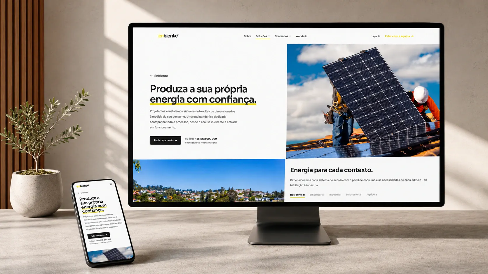 A página inicial do site reconstruído da Enbiente, num monitor e num telemóvel sobre uma secretária: título «Produza a sua própria energia com confiança», fotografia de instaladores a montar painéis solares e o menu de navegação.